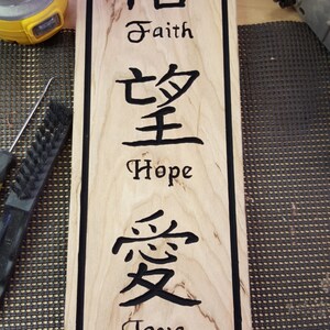 May include: A wooden sign with black lettering. The sign has the Chinese characters for faith, hope, and love, with the English words below each character. The sign is on a wooden surface.