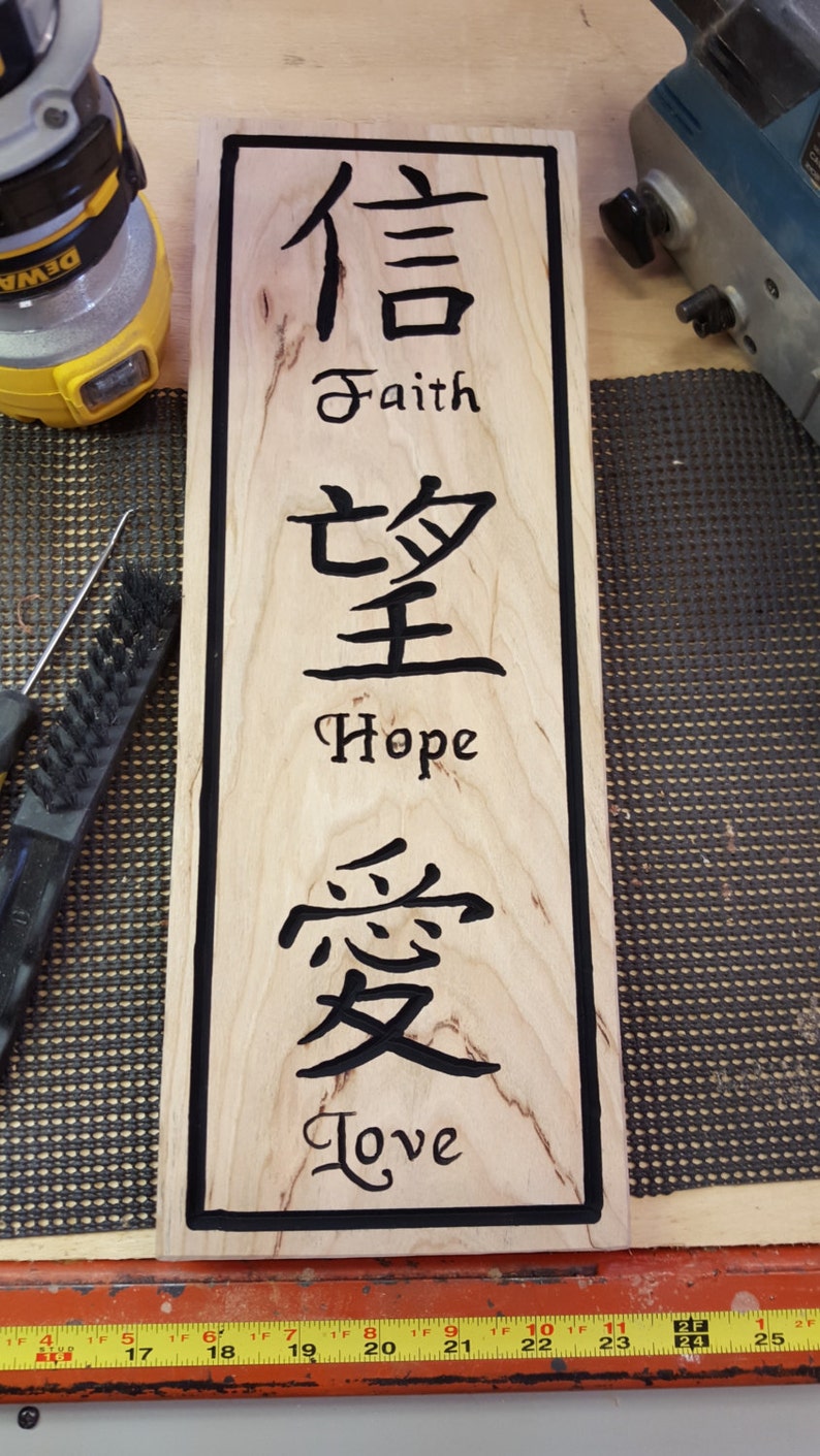 May include: A wooden sign with black lettering. The sign has the Chinese characters for faith, hope, and love, with the English words below each character. The sign is on a wooden surface.