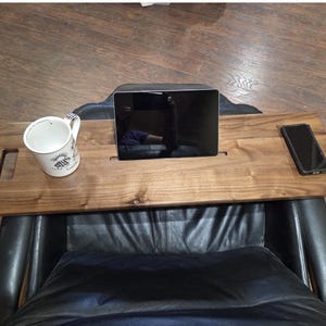 May include: A rectangular brown wooden lap desk tray featuring a smooth finish, two side handles, a central slot for a tablet, and a recessed rectangular area on the right side for a smartphone. The tray is positioned across a black chair.