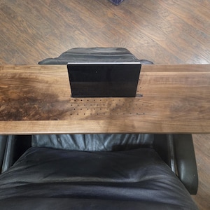 May include: A rectangular dark brown wooden lap desk featuring a central slot for holding a tablet or smartphone, a series of small ventilation holes, and a cutout handle on the right side. The surface displays a natural wood grain pattern.
