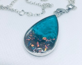 May include: A silver necklace with a teardrop pendant. The pendant features a teal blue resin top and a dark, textured bottom with flecks of orange and yellow. The necklace is on a white background.