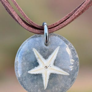 Pode incluir: Um colar com pingente redondo com uma estrela-do-mar branca encapsulada em resina transparente texturizada. O pingente é suspenso por um cordão de camurça marrom. A estrela-do-mar tem uma superfície detalhada e texturizada.