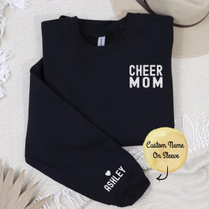 Könnte beinhalten: Schwarzer Sweatshirt mit dem Text "CHEER MOM" auf der Vorderseite und einem Herz mit dem Namen "ASHLEY" auf dem Ärmel.