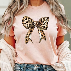 Puede incluir: Una camiseta rosa con un gran lazo de estampado de leopardo en la parte delantera. El lazo est&aacute; dibujado en un estilo realista con manchas marrones y negras.