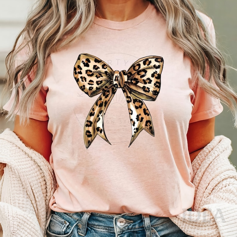 Puede incluir: Una camiseta rosa con un gran lazo de estampado de leopardo en la parte delantera. El lazo est&aacute; dibujado en un estilo realista con manchas marrones y negras.