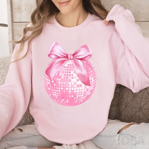 May include: Pink sweatshirt featuring a pink disco ball with a pink bow.