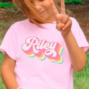 May include: A pink t-shirt with a retro rainbow design featuring the name "Riley" in white.