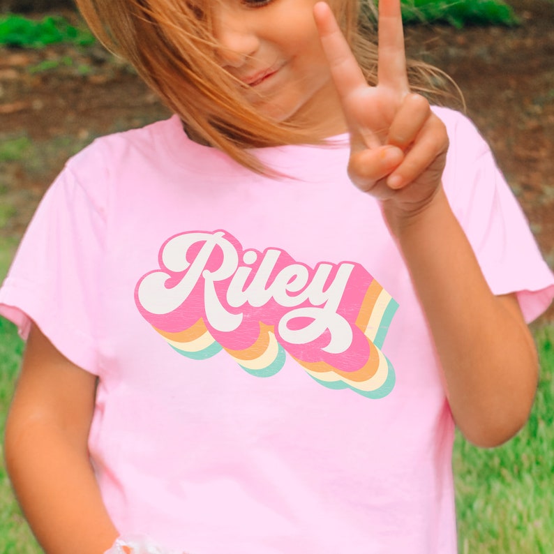 May include: A pink t-shirt with a retro rainbow design featuring the name "Riley" in white.
