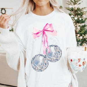 May include: White t-shirt with a pink bow and two silver disco ball ornaments hanging from it. The text "Kidspa" and "Work" are visible on the shirt.