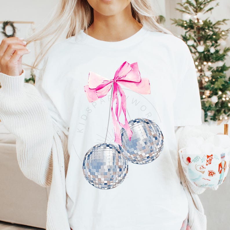 May include: White t-shirt with a pink bow and two silver disco ball ornaments hanging from it. The text "Kidspa" and "Work" are visible on the shirt.