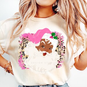 May include: A white t-shirt with a pink and black Santa Claus graphic. The Santa Claus is wearing a pink hat and has a white beard. The graphic is surrounded by a pink and gold leopard print design.