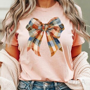 Pu&ograve; includere: Una maglietta rosa con un grande fiocco in stile scozzese ad acquerello in tonalit&agrave; di blu, arancio e marrone.