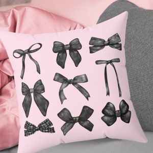 May include: A pink throw pillow with black and white illustrations of bows. The pillow has a soft, pink fabric and a white background.