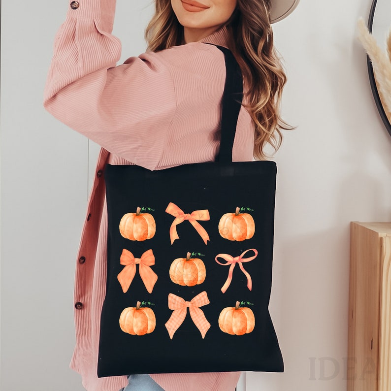 May include: Black canvas tote bag featuring a design of orange pumpkins and orange bows. The pumpkins have green stems and leaves.
