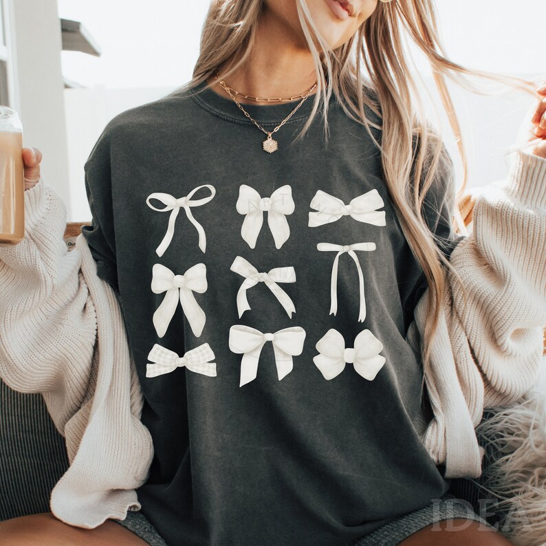 May include: A dark gray t-shirt with a white graphic of various bow designs. The shirt has a relaxed fit and a crew neck.