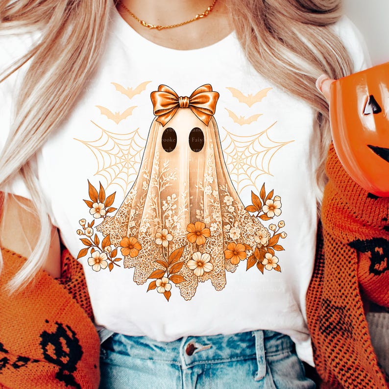 May include: White t-shirt featuring a Halloween ghost design. The ghost is draped in a lace-like fabric with a bow, surrounded by flowers, bats, and a spiderweb. The ghost has two black oval eyes. The text on the ghost is blurry.