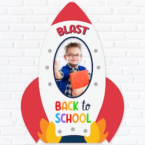 May include: A white rocket-shaped cardboard cutout with a red border and a photo frame in the center. The frame has a boy wearing a plaid shirt and glasses looking at the camera. The text "Blast" is at the top of the cutout and "Back to School" is at the bottom.