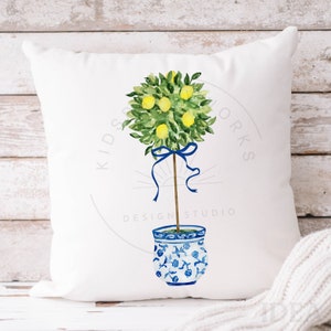 Pode incluir: Uma almofada branca com uma ilustra&ccedil;&atilde;o em aquarela de uma &aacute;rvore de lim&atilde;o em um vaso com padr&atilde;o azul e branco. A &aacute;rvore est&aacute; amarrada com uma fita azul e tem folhas verdes e lim&otilde;es amarelos. O texto "Kids Design Studio Works" est&aacute; impresso na almofada.
