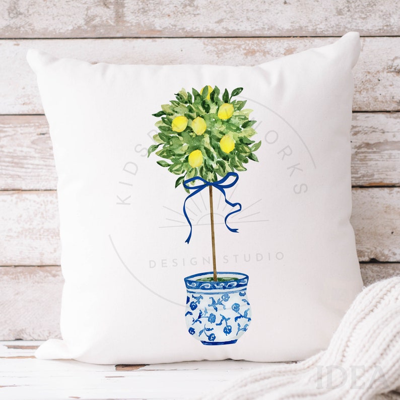 Pode incluir: Uma almofada branca com uma ilustra&ccedil;&atilde;o em aquarela de uma &aacute;rvore de lim&atilde;o em um vaso com padr&atilde;o azul e branco. A &aacute;rvore est&aacute; amarrada com uma fita azul e tem folhas verdes e lim&otilde;es amarelos. O texto "Kids Design Studio Works" est&aacute; impresso na almofada.