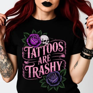 May include: Black t-shirt featuring a graphic design with the text "Tattoos Are Trashy". The design includes purple roses, green leaves, and a white skull. The text is in a pink, stylized font.