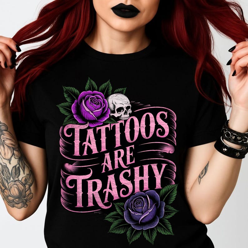 May include: Black t-shirt featuring a graphic design with the text "Tattoos Are Trashy". The design includes purple roses, green leaves, and a white skull. The text is in a pink, stylized font.