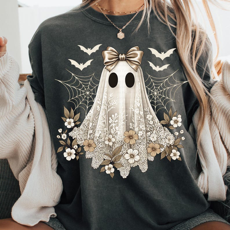 May include: Dark gray sweatshirt featuring a ghost design with a bow, floral accents, and spiderwebs. The ghost has two black oval eyes and is draped in a white lace-like fabric. Bats are also featured in the design, creating a Halloween theme.