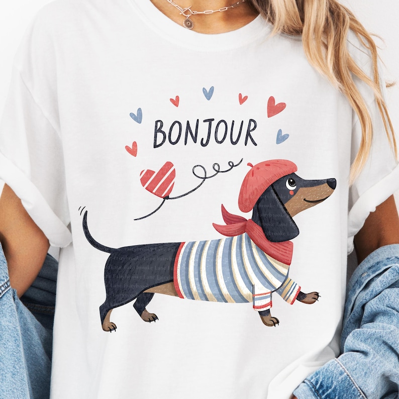 May include: White t-shirt featuring a cartoon dachshund wearing a red beret and scarf, and a blue and white striped shirt. The word "Bonjour" is above the dog, with heart and arrow graphics.