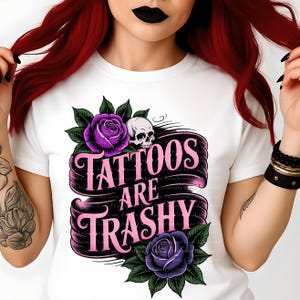 May include: White t-shirt featuring a graphic design with the text "Tattoos Are Trashy" in pink, accompanied by purple roses, green leaves, and a skull. The design is centered on the front of the shirt.