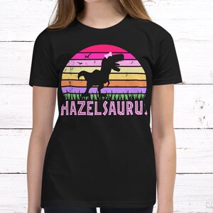May include: Black t-shirt with a pink, purple, orange, and green retro sunset graphic featuring a dinosaur with a pink bow. The text "HAZELSAURUS" is printed in pink below the graphic.
