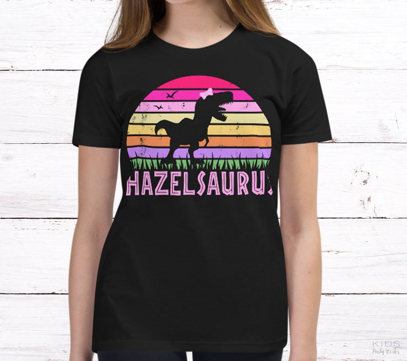 May include: Black t-shirt with a pink, purple, orange, and green retro sunset graphic featuring a dinosaur with a pink bow. The text "HAZELSAURUS" is printed in pink below the graphic.