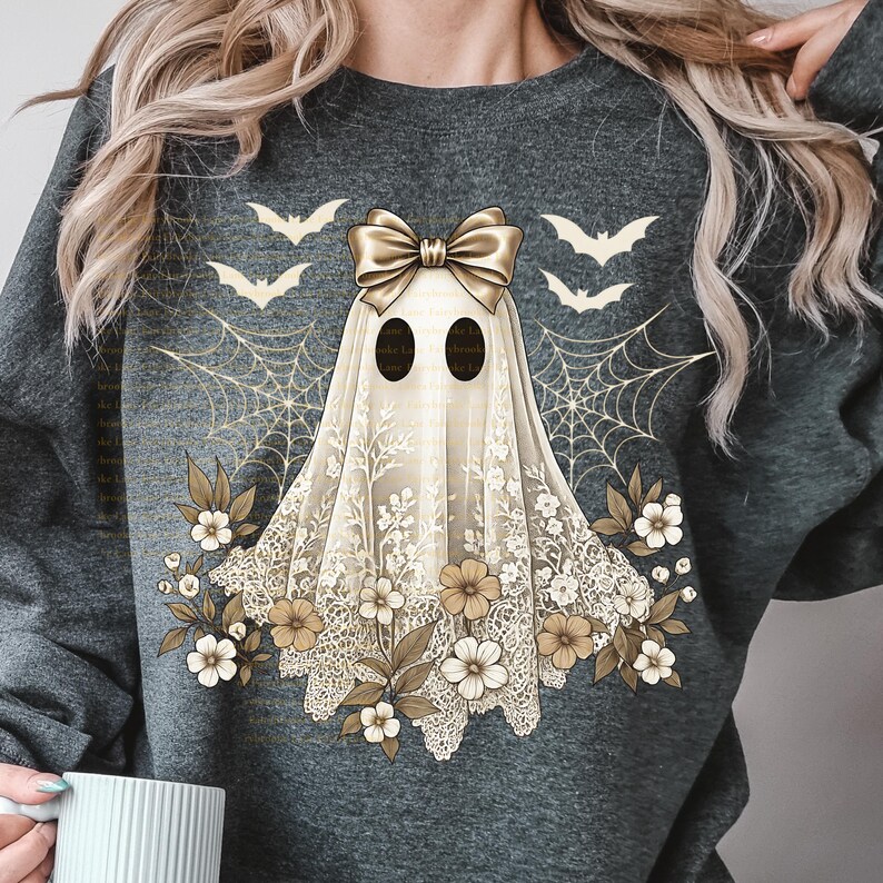 May include: A gray sweatshirt featuring a ghost design with a gold bow, lace detailing, and floral accents. The ghost has two black oval eyes and is surrounded by bats and spiderwebs. A Halloween-themed graphic.