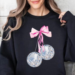 May include: Black sweatshirt with a pink bow and two silver disco balls hanging from it.