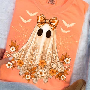 May include: An orange t-shirt featuring a ghost design with a bow, lace, flowers, and bats. The ghost has large black eyes and is draped in a white lace sheet. The design includes a spider web and autumn-colored flowers.