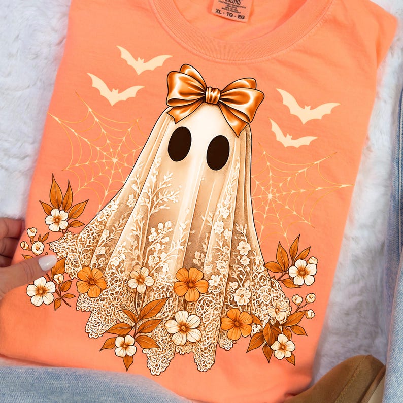 May include: An orange t-shirt featuring a ghost design with a bow, lace, flowers, and bats. The ghost has large black eyes and is draped in a white lace sheet. The design includes a spider web and autumn-colored flowers.