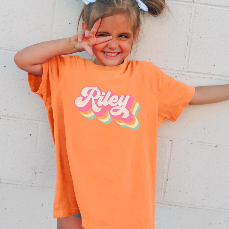 May include: A young person wearing an orange t-shirt with a retro-style graphic that says "Riley" in pink letters with rainbow stripes.