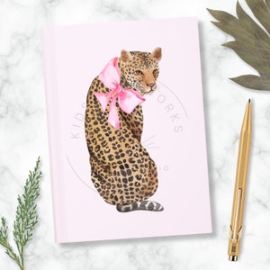 Puede incluir: Una libreta rosa con una ilustraci&oacute;n de acuarela de un leopardo con un lazo rosa. La libreta tiene el texto "Kids Works Studio" impreso en la portada.