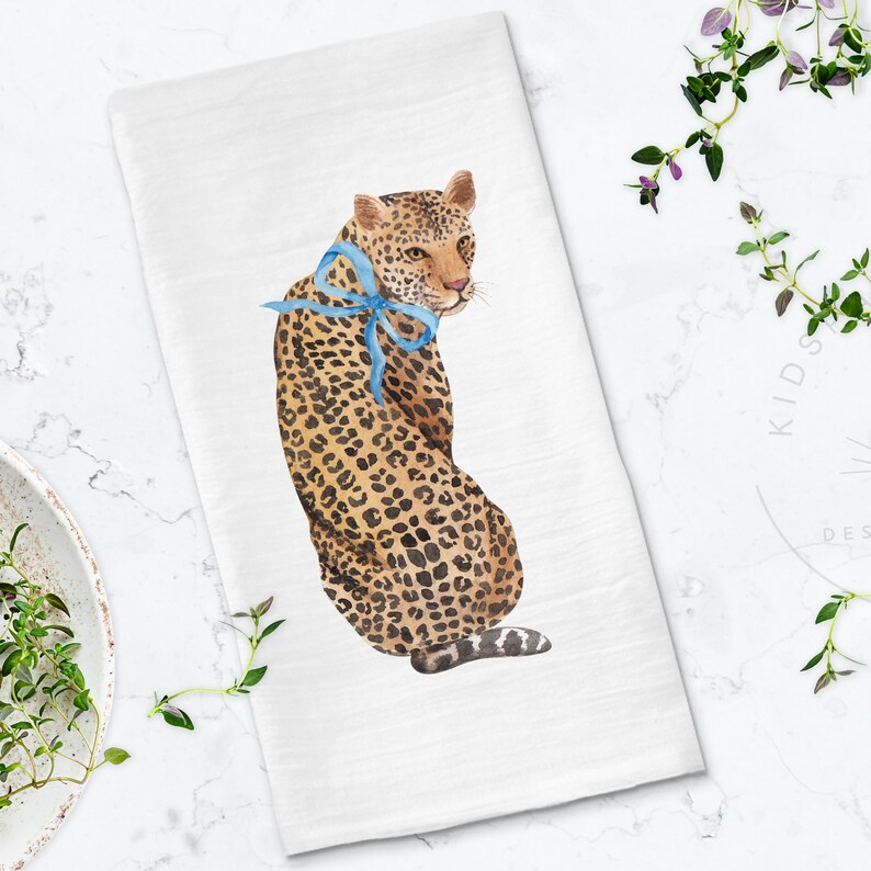 Puede incluir: Una toalla de cocina blanca con una ilustraci&oacute;n de acuarela de un leopardo con un lazo azul. El leopardo est&aacute; de espaldas al espectador y tiene un patr&oacute;n de manchas.