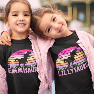 May include: Two girls wearing black t-shirts with a retro sunset graphic and a dinosaur silhouette. The shirts have the names "EMMISAUR" and "LILLYSAUR" printed on them.