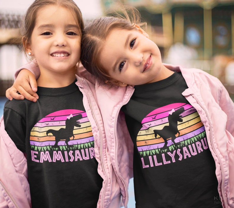 May include: Two girls wearing black t-shirts with a retro sunset graphic and a dinosaur silhouette. The shirts have the names "EMMISAUR" and "LILLYSAUR" printed on them.