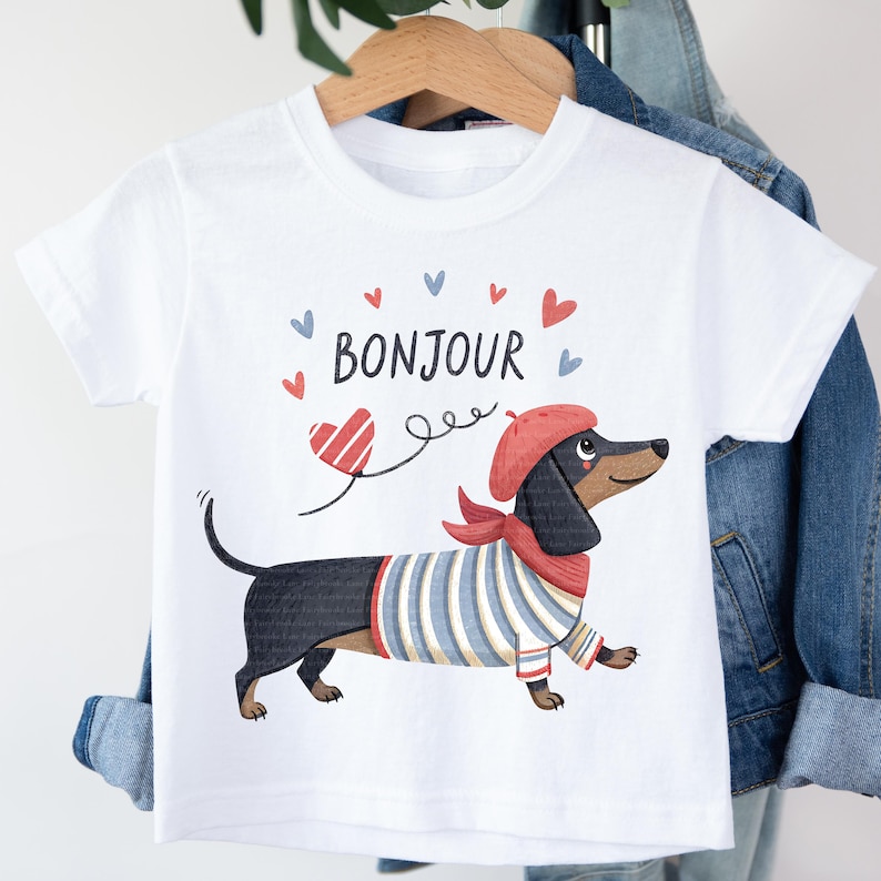 May include: White t-shirt featuring a cartoon dachshund wearing a red beret and scarf, and a blue and white striped shirt. The word "BONJOUR" is above the dog, with red and blue hearts. The shirt is hanging on a wooden hanger.