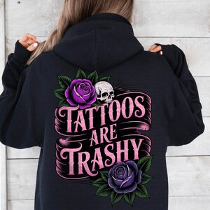 May include: Black hoodie with a graphic design on the back. The design features a skull, purple roses, and the text "Tattoos Are Trashy" in pink lettering. The hoodie is a dark color.