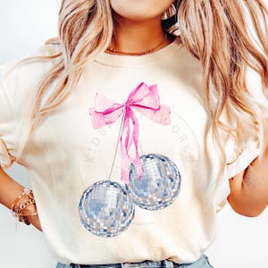 May include: A white t-shirt with a pink bow and two silver disco balls hanging from it. The text "Kids Works" is visible on the shirt.