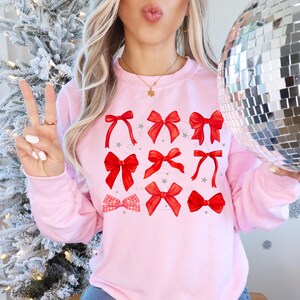 Puede incluir: Una sudadera rosa con un dise&ntilde;o festivo de lazos rojos. Los lazos est&aacute;n dispuestos en una cuadr&iacute;cula y est&aacute;n rodeados de peque&ntilde;as estrellas blancas.