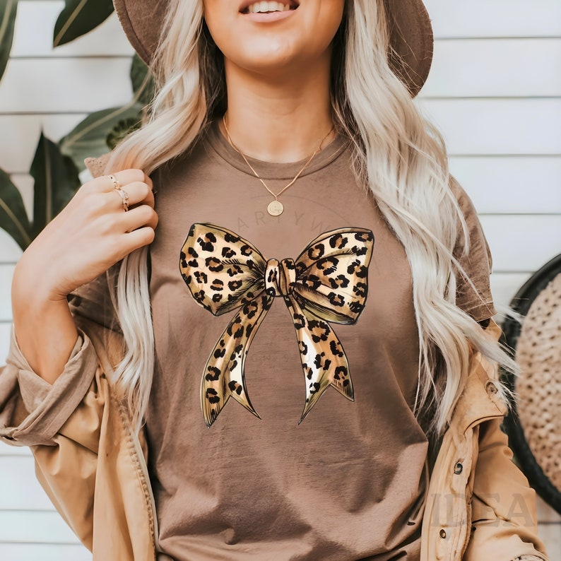 Puede incluir: Una camiseta marr&oacute;n con un gran lazo de estampado de leopardo en la parte delantera. El lazo est&aacute; hecho de tela marr&oacute;n y dorada con un contorno negro.