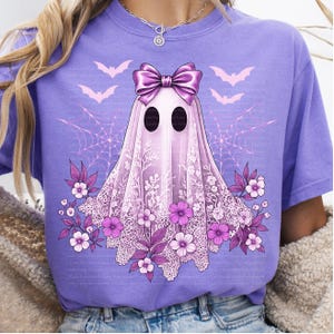 May include: A lilac-colored t-shirt featuring a ghost design with a lace veil, bow, and floral accents. The ghost has black oval eyes and is surrounded by bats and spiderwebs. The design is in shades of purple and white.