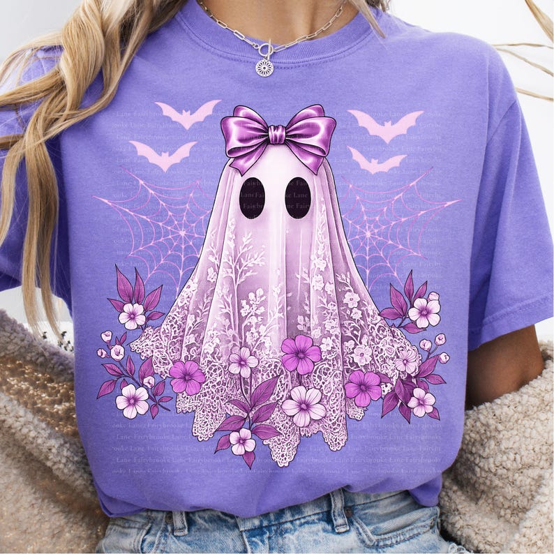 May include: A lilac-colored t-shirt featuring a ghost design with a lace veil, bow, and floral accents. The ghost has black oval eyes and is surrounded by bats and spiderwebs. The design is in shades of purple and white.
