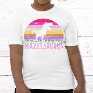 May include: White t-shirt with a pink, purple, and orange sunset graphic featuring a dinosaur with a flower on its head. The text "HAZELSAURUS" is printed in pink below the graphic.