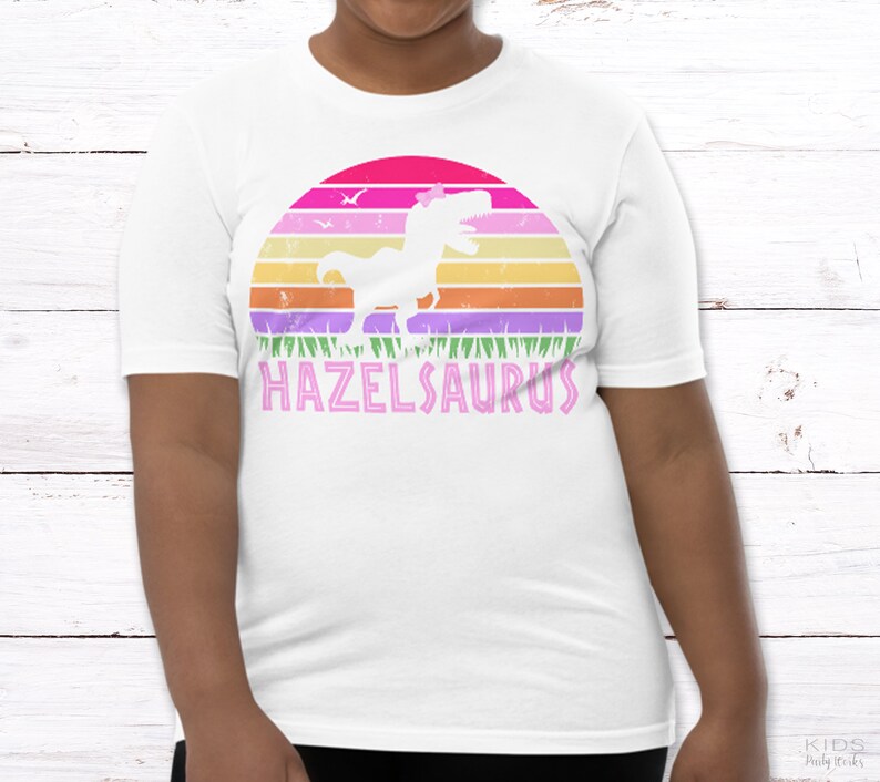 May include: White t-shirt with a pink, purple, and orange sunset graphic featuring a dinosaur with a flower on its head. The text "HAZELSAURUS" is printed in pink below the graphic.