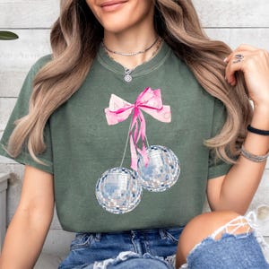 May include: A green t-shirt with a pink bow and two silver disco ball ornaments hanging from it.