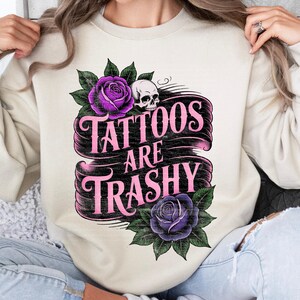 May include: Cream-colored sweatshirt with the text "Tattoos Are Trashy" in pink script on a black banner. The design includes purple roses, green leaves, and a small skull.